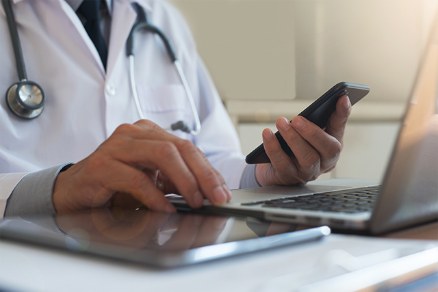 a man in a white coat sits at a desk on his phone and laptop demonstrating cyber analytics careers in healthcare