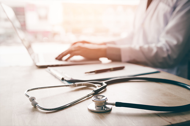 a person in a white lab coat sits at a desk with a laptop and stethoscope facing cybersecurity challenges in healthcare