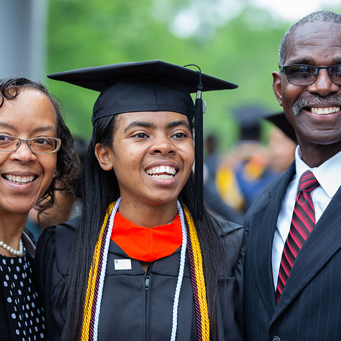 A female graduating Capitol Tech student in cap and gown standing for a picture with her mother and father.