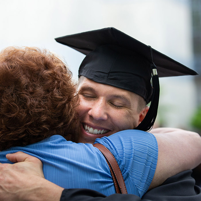 A graduating Capitol Tech student in cap and gown, hugging their parent on graduation day.
