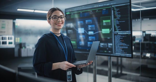 a woman in the STEM field holds a laptop surrounded by monitors