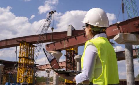 woman in a hard hat with a laptop supervises as a bridge is built demonstrating technological advancements in construction for critical infrastructure